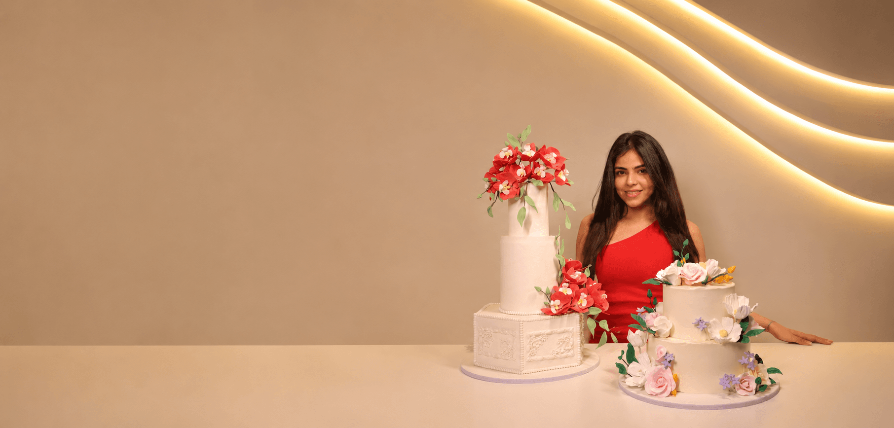 An Amari Bakehouse baker beside a tall red-orchid tiered cake and a pastel floral wedding cake