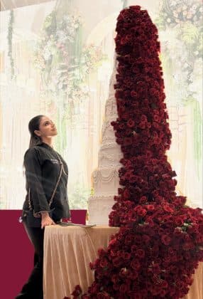 An Amari Bakehouse chef beside a hand-piped tiered cake with a cascading red rose garland